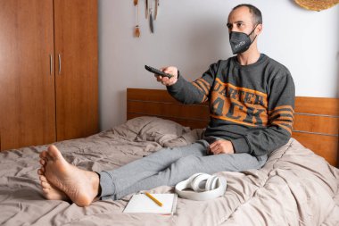 Kulaklıklı, cep telefonu ve yüz maskeli bir adam, yatağında oturuyor ve odasına kapatılmış.