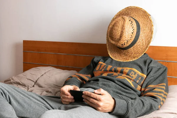 Άντρας με ακουστικά, κινητό τηλέφωνο και μάσκα προσώπου, κάθεται στο κρεβάτι του και περιορίζεται στο δωμάτιό του — Φωτογραφία Αρχείου
