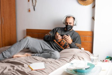 Kulaklıklı, cep telefonu ve yüz maskeli bir adam, yatağında oturuyor ve odasına kapatılmış.