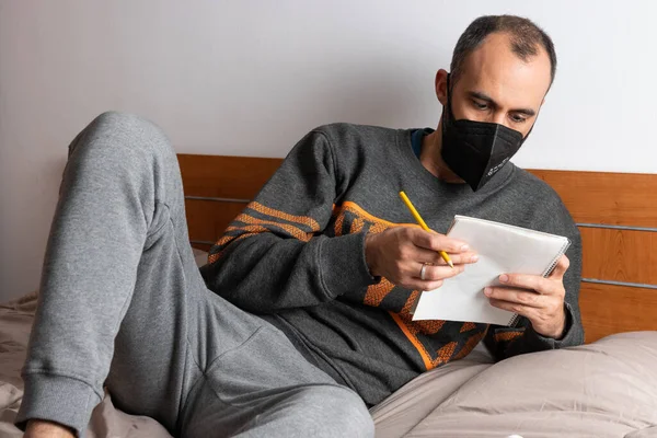 Человек в наушниках, мобильном телефоне и маске, сидящий на кровати и запертый в своей комнате — стоковое фото