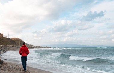 Bulutlu bir günde sahilde yürüyen adam çalkantılı suyla