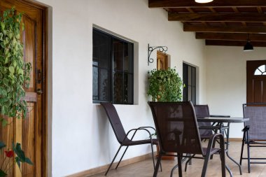 Ahşap zemin üzerinde sandalyeler ve masa olan bir veranda. Kapı, pencere ve bitkilerle ön cephe