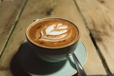 Tabağı ve kaşığı tahtadan bir masada duran fincan. Yaprak çizimli kahve ya da kapuçino.