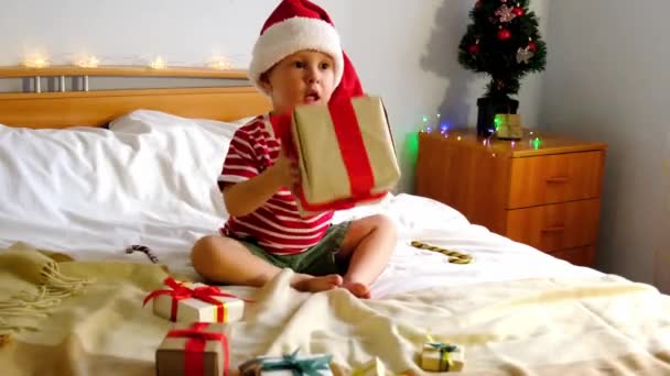 Un niño, sentado en la cama, mirando y sacudiendo cajas de regalos de Navidad — Vídeos de Stock