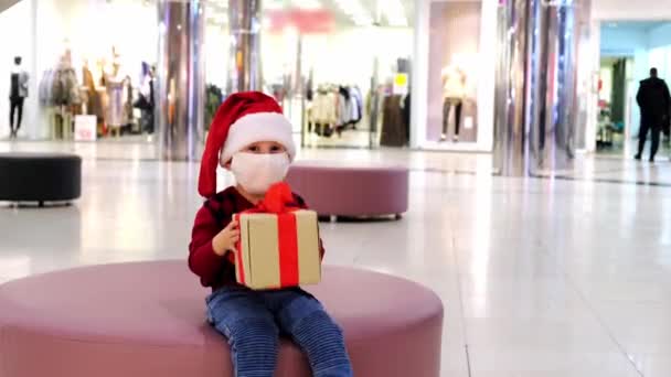Ein Kleinkind Mit Weihnachtsmütze Und Medizinischer Maske Sitzt Auf Einem — Stockvideo