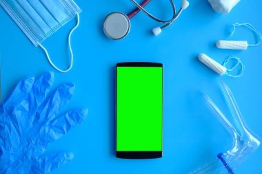 Tıbbi ekipman stetoskop, jinekolojik ayna eldiveni ve maske ve mavi arka planda yeşil ekranlı telefon..