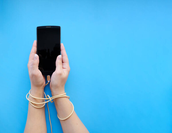 Крупный план сверху женских рук, соединенных проводным кабелем со смартфоном, на синем фоне. 