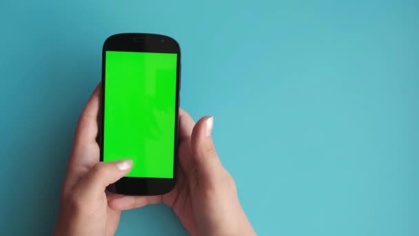 Mano femenina utiliza un teléfono con una pantalla verde sobre un fondo azul uniforme, — Vídeos de Stock