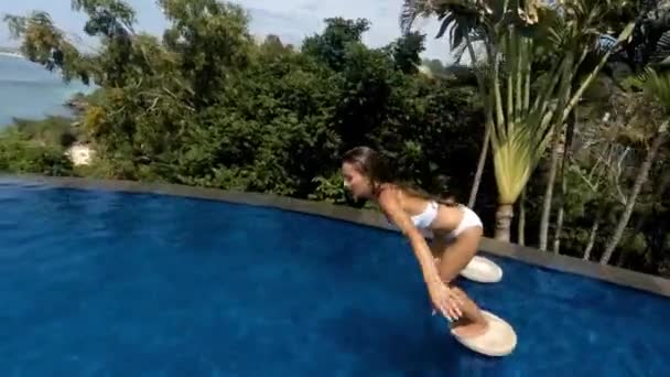 Eine aktive junge Frau im Badeanzug taucht ins Becken und spritzt glasiges Wasser. Mädchen springt in den Pool, während sie sich in einem tropischen Resort vor dem Hintergrund grüner Palmen und des Ozeans entspannen — Stockvideo