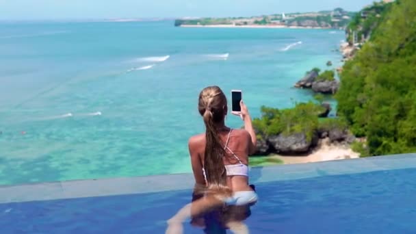 En ung og smuk pige i en udendørs pool på en tropisk ø tager en smartphone kamera landskab. Kvinde svømning i poolen med udsigt over havet – Stock-video