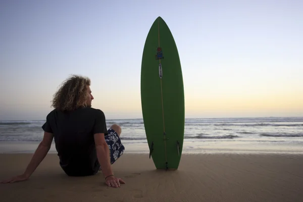 Gün batımını izlerken onun surfboard ile sahilde oturan genç bir adam. — Stok fotoğraf