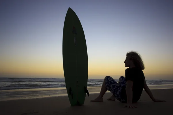 Un joven sentado en la playa con su tabla de surf mirando el atardecer . —  Fotos de Stock