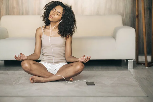 精神衛生の概念。自宅で瞑想を練習若い女性,イージーシートポーズに座って,彼女の目を閉じて,電話でオーディオ瞑想を聞きます ロイヤリティフリーのストック写真