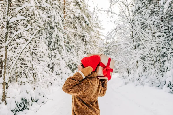 Musim dingin konsep belanja Natal. Seorang wanita muda memegang kotak hadiah besar dengan pita merah dan berjalan maju di hutan musim dingin. — Stok Foto