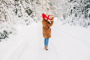 Kahverengi bir kürk manto ve kırmızı bir şapka giyen genç bir kadın büyük bir hediye kutusu ve kırmızı bir kurdele tutuyor ve kar ormanlarında ileri doğru yürüyor. Noel hediyesi ve alışveriş konsepti. Arka plan