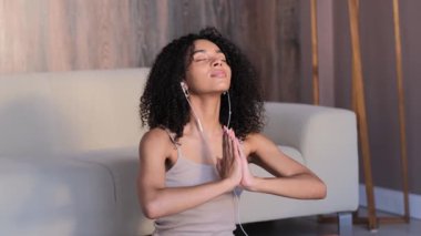 African woman Meditating, doing Yoga at home in wooden interior. Curly black woman listens to meditation with headphones and relaxes.