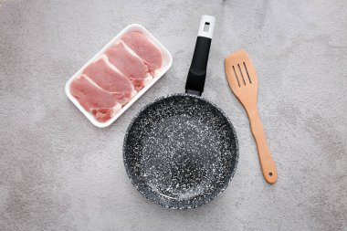 Black marble frying pan with raw meat and wooden spatula on grey table background, top view