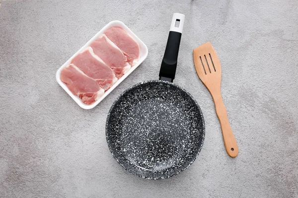 Black marble frying pan with raw meat and wooden spatula on grey table background, top view