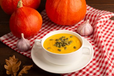 Balkabağı çorbası kremalı ve kabak tohumlu koyu ahşap arka planda. Arka planda kırmızı kumaşlı balkabağı ve sarımsaklı. Boş alan, yakın plan