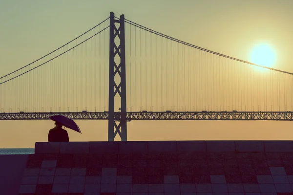 背景の橋と夕日に座って傘を持つ女性, — ストック写真