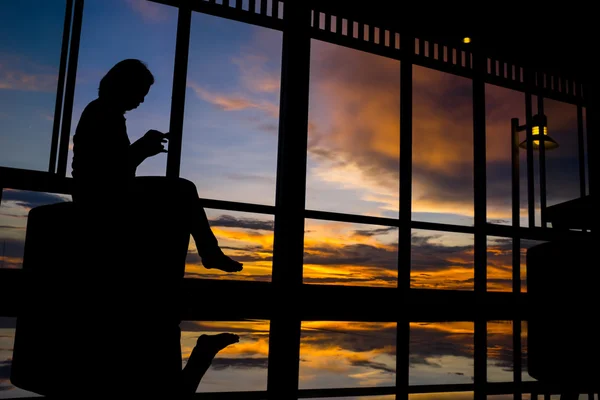 Girl playing smart phone by window at sunset with twilight sky — Stock Photo, Image