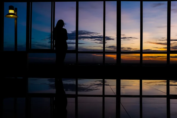 Dziewczyna przy użyciu inteligentny telefon przez okno na zachód słońca, Zmierzch niebo — Zdjęcie stockowe