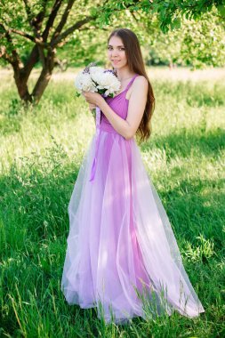 Portrait of a young attractive bridesmaid girl in a delicate purple dress with long hair in a spring apple garden among the greenery. Bouquet of peonies in hands. High quality photo.