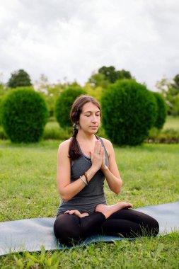 Front view of a woman instructor sitting on yoga mat in lotus position outside in city park. Namaste mudra, seated yoga asana, lotus pose, lotus posture, padmasana, cross legged sitting posture.