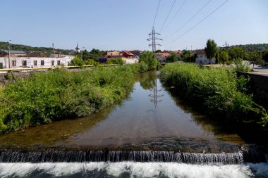 Hunedoara, Hunedoara County - July 22, 2022: View of the Cerna river passing through the city of Hunedoara, Transylvania, Romania. Power poles are reflected in the water.