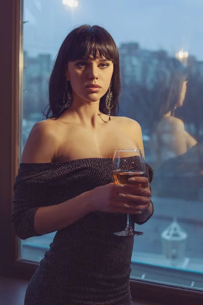 美丽的女人站在黄昏的窗前 手里拿着一杯葡萄酒 这个女人的肩膀是开着的 看起来很性感 — 图库照片