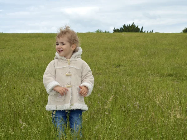 Young girl walking in field. She is wearing a cream coat and blue jeans. She has blonde curly hair, blowing in the wind. She is standing in a meadow of long grass. — Stock Photo, Image