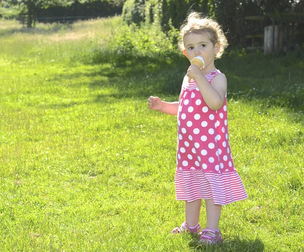 Pretty young girl eating ice cream. She is wearing a pink and white dress, with the sun behind her (backlit). She has blonde curly hair and is looking towards the camera. — Stock Photo, Image