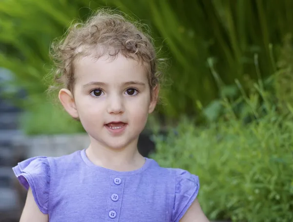 Criança no jardim. Uma menina bonita, criança, usando um top lilás roxo. Cabeça e ombros atingidos, com fundo verde de jardim. Ela tem cabelo encaracolado loiro e uma boa expressão . — Fotografia de Stock