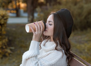 Beyaz kazaklı ve siyah bereli genç bir kız sonbaharda bankta kahve içer..