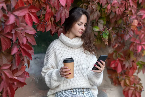 A girl in a white jumper looks at the phone and holds a glass.