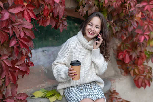 A girl in a white jumper is talking on the phone and holding a glass.