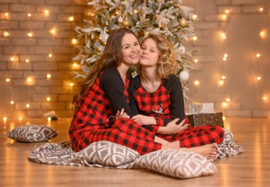 Anne ve kızı kırmızı pijamalarıyla stüdyoda yerde, yatak örtüsünün üzerinde sarılıyorlar..