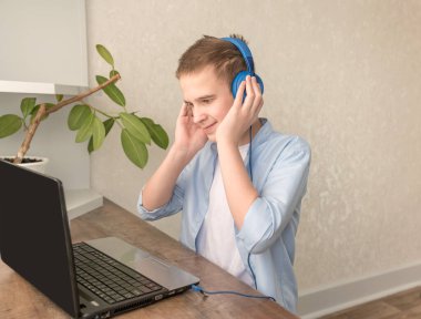 A teenager in a blue shirt at home in headphones at the table in front of a laptop. Listening to music.