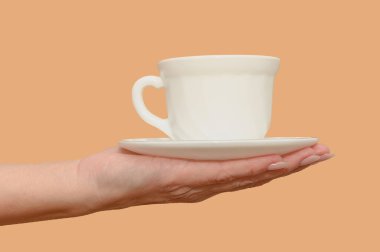 A female hand holds a white cup and saucer. Minimalism. Close-up. On a beige background.