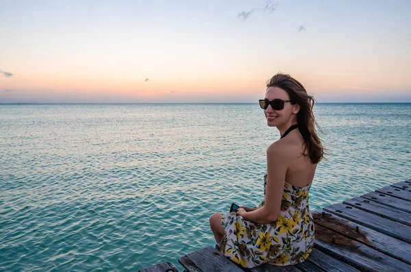Jovem mulher sentada e sorrindo no molhe à beira-mar ao pôr do sol bonito — Fotografia de Stock
