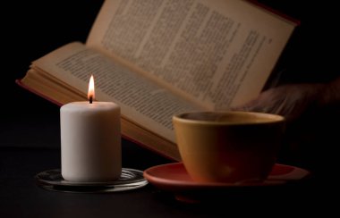 Alevli bir mum, dumanlı bir fincan çay ve arka planda bir okuma kitabı. (muma odaklan).