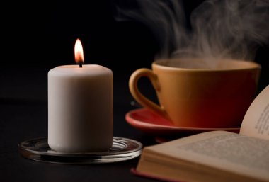 Alevli bir mum, dumanlı bir fincan çay ve arka planda bir okuma kitabı. (muma odaklan).