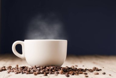 Sıcak buharlı bir fincan kahve, ahşap masada kavrulmuş kahve çekirdekleri, mavi arka planda..