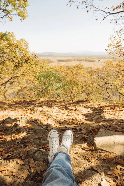展望台の秋の森を背景にジーンズと白いスニーカーを履いた女性の足 ハイキングをしたり山に登ったり景色を眺めたり 選択的焦点 ストック写真