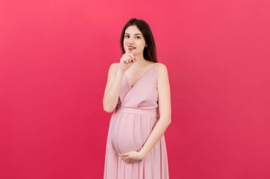 Genç, güzel, esmer, hamile bir kadın izole edilmiş renkli bir arka planda bebek bekliyor ve parmakları dudaklarında sessiz olmak istiyor. Annelik Sessizliği ve gizli konsept.