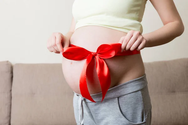 Bauch Einer Schwangeren Frau Mit Roter Schleife Vor Heimischem Hintergrund — Stockfoto