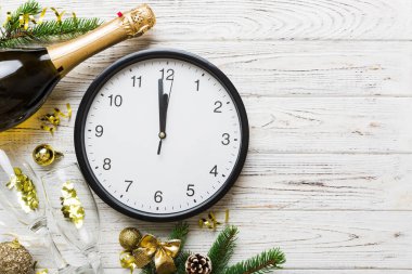 New year celebration concept with a bottle of champagne and two glasses toasting. Christmas gift box, alarm clock and fir tree branch on colored table. Top view Copy space.