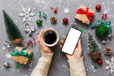 Bir kadının elinde sıcak bir fincan kahveyle akıllı bir telefonu tutması. Renkli arka planda Noel süslemesi.