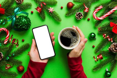Bir kadının elinde sıcak bir fincan kahveyle akıllı bir telefonu tutması. Renkli arka planda Noel süslemesi.