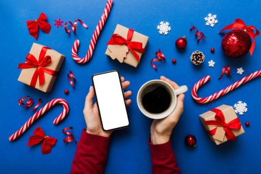 Bir kadının elinde sıcak bir fincan kahveyle akıllı bir telefonu tutması. Renkli arka planda Noel süslemesi.